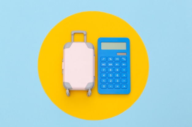 Despesas de viagem. Mini mala de viagem de plástico com calculadora em fundo azul com círculo amarelo. Vista do topo. Postura plana