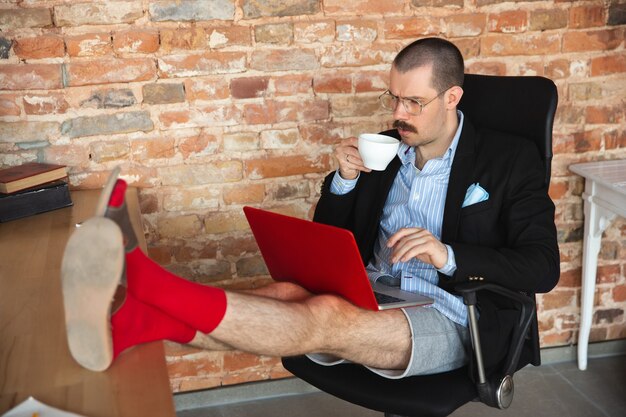 Desplazamiento. Hombre joven sin pantalones pero con chaqueta trabajando en una computadora, computadora portátil. Oficina remota durante el coronavirus, divertida y cómoda en calzoncillos. Aislamiento, cuarentena, humor, concepto empresarial.