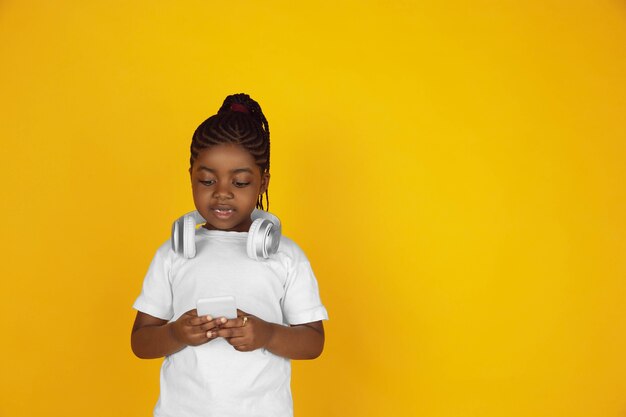 Desplazamiento del teléfono, escuchar música. Retrato de niña afroamericana sobre fondo amarillo de estudio. Niño alegre y hermoso. Concepto de emociones humanas, expresión, ventas, publicidad. Copyspace. Se ve lindo.