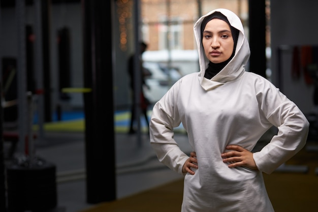 Desportista árabe está posando no ginásio após o treino, retrato de uma senhora muçulmana em hijab branco posando, olhando para a câmera.