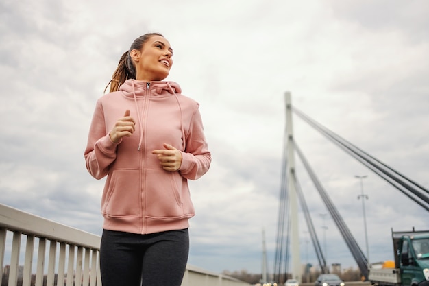 Desportista de ajuste atraente sorridente com agasalho, correndo na ponte em tempo nublado.