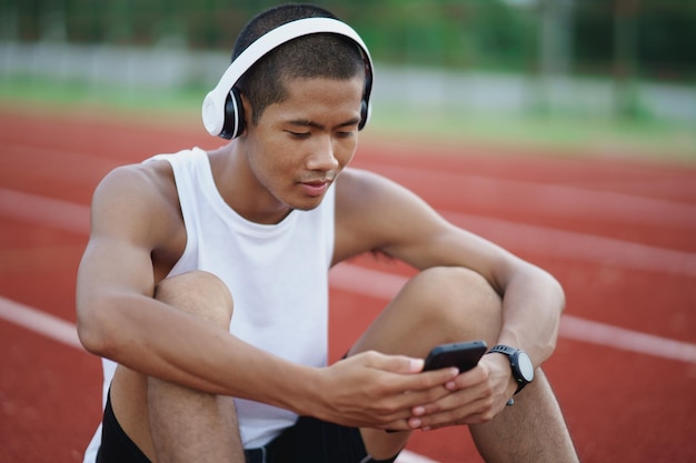 Desportista de homem asiático em forma de corpo usando fone de ouvido branco e usando o celular para ouvir música enquanto descansa ou antes de correr se exercitando no estádio de pista Conceito de corrida saudável esportiva