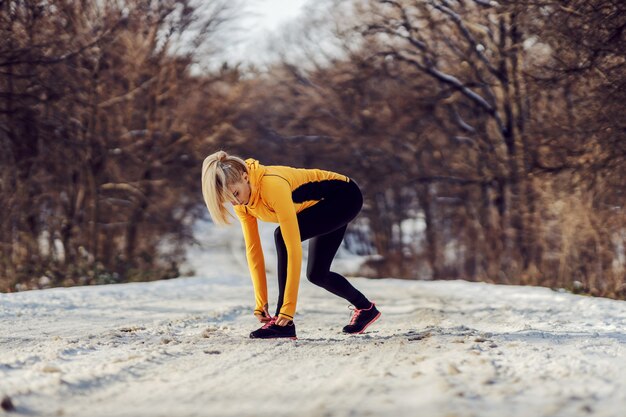 Desportista de pé no caminho nevado na natureza no inverno e amarrando o cadarço. Roupa esportiva, fitness de inverno, vida saudável