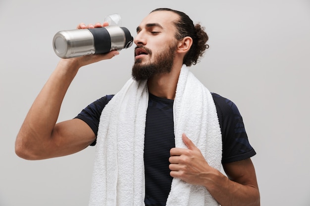 Desportista musculoso sério com toalha sobre o pescoço bebendo água após treino isolado na parede branca