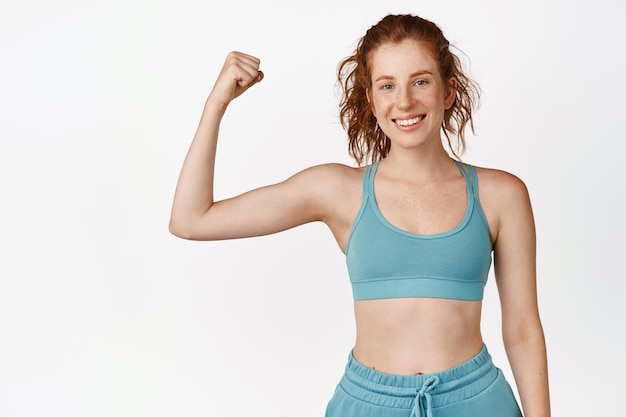 Desportista ruiva ativa e saudável flexionando bíceps e sorrindo treino de garota fitness mostrando os músculos do braço usando sutiã esportivo em pé sobre fundo branco