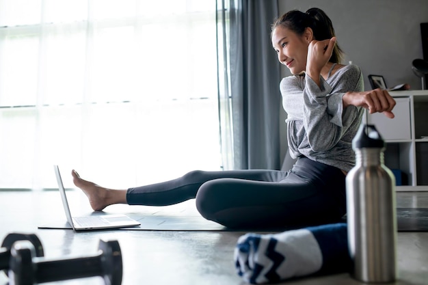 Desportiva jovem asiática se exercitando em casa assistindo a vídeos de fitness na internet ou tendo aula de fitness on-line usando o espaço de cópia interior da sala de estar do laptop