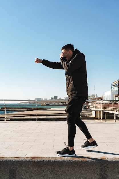 desportivo homem 20 anos com fato de treino preto a lutar boxe e a dar soco durante o treino matinal à beira-mar