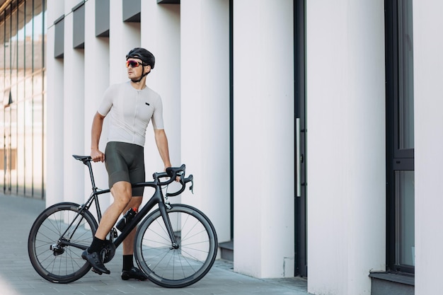 Desportivo jovem no capacete activewear e óculos de pé com bicicleta perto de um edifício moderno e olhando de lado cara caucasiano, passando tempo livre para treinar ao ar livre