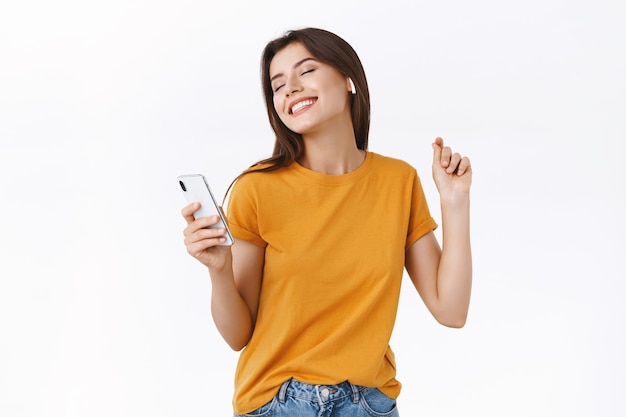 Despreocupada, terna e elegante mulher moderna em uma camiseta amarela curtindo a batida incrível em novos fones de ouvido sem fio, dançando com as mãos levantadas e pose relaxada, sorrindo de perto segurando um smartphone