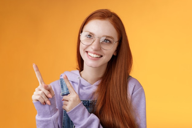 Despreocupado carismático feliz bobo jovem ruiva bonita sardas olhos azuis de óculos desfrutar de contemplar a exposição divertida apontando para a esquerda indicando, fundo laranja.