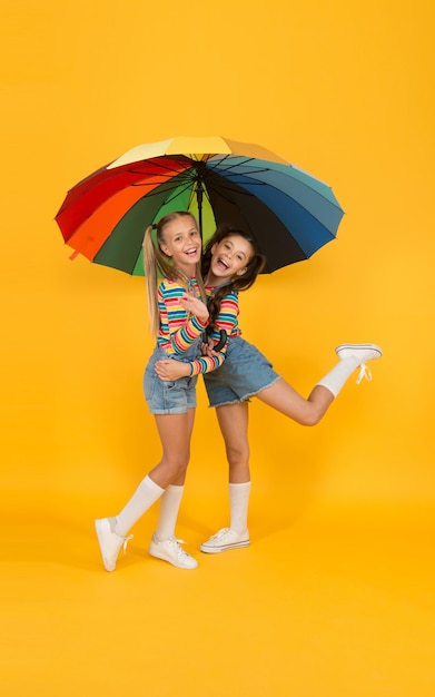 Despreocupados y felices, dos niños felices, niños de fondo amarillo, disfrutan de la moda infantil de otoño lluvioso, sintiéndose seguros y cómodos, buen humor en cualquier clima, niña pequeña bajo un paraguas colorido