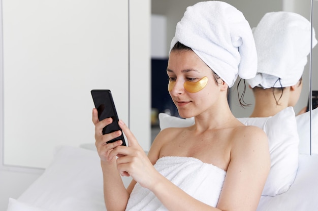 Después de una ducha, la niña está envuelta en una toalla y usa parches cosméticos para la piel debajo de los ojos Mira el teléfono celular Procedimientos cosméticos en casa Preparándose para una cita