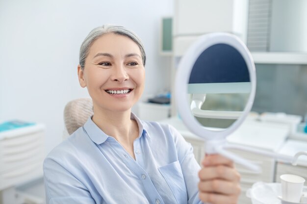 Después de la restauración de los dientes. Mujer sentada en la oficina de dentistas con un espejo en las manos