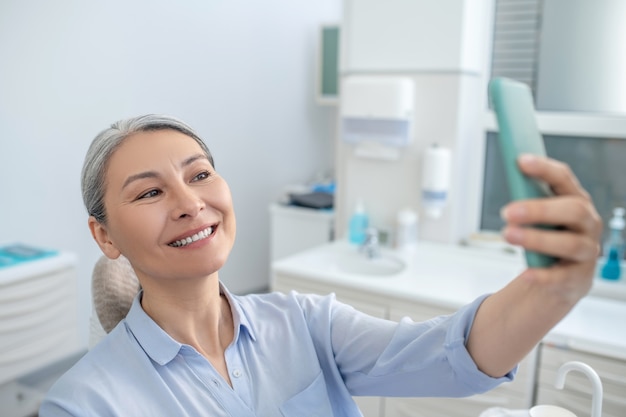 Después de la restauración de los dientes. Sonriente mujer bastante madura con un teléfono inteligente en la oficina de dentistas