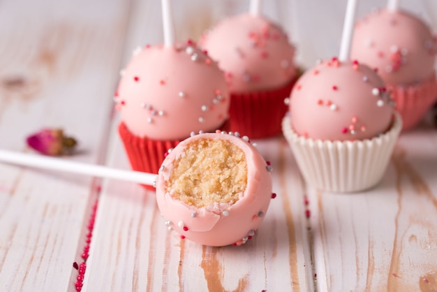 Dessert auf einem Stock in rosa Creme mit einem Streusel auf einem hölzernen Hintergrund. Cake Pops zwischen ganzen Desserts geschnitten.