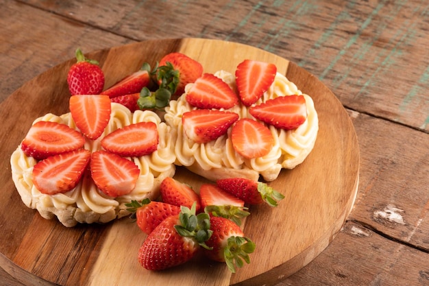 Dessert aus zwei Waffeln mit weißer Schokoladencreme und Erdbeerstücken auf einem runden Holzbrett und geschnittenen Erdbeeren auf einem Holztisch in Nahaufnahme