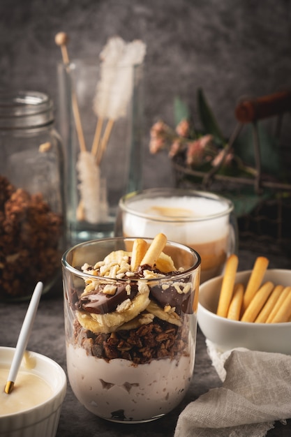 Dessert in einem Glas Joghurt, Müsli, Banane und Schokolade mit Keksen, Cappuccino.