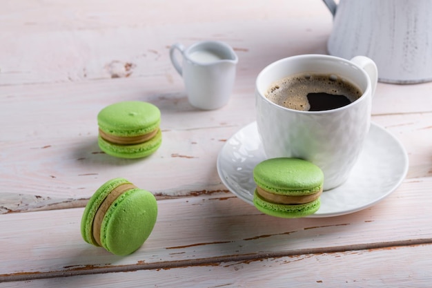 Dessert mit französischen Makronen Drei grüne Makronen und eine weiße Tasse Kaffee auf einem weißen Holztisch