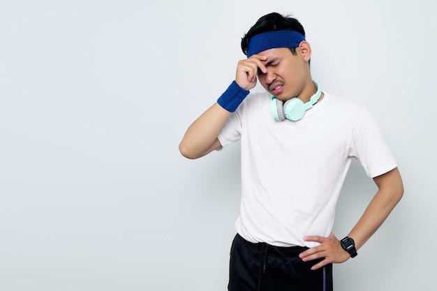 Destacado joven deportista asiático en diadema azul y camiseta blanca de ropa deportiva con auriculares tocando sus sienes y con dolor de cabeza aislado sobre fondo blanco Concepto de deporte de entrenamiento