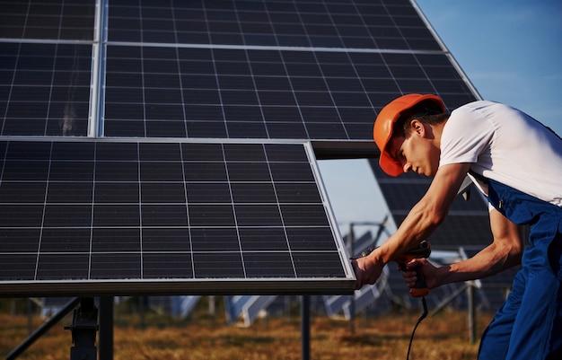 Con destornillador inalámbrico. Trabajador de sexo masculino en uniforme azul al aire libre con baterías solares en un día soleado.