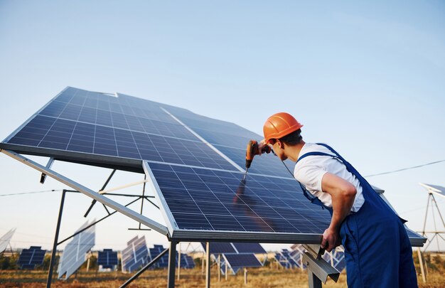 Con destornillador inalámbrico. Trabajador de sexo masculino en uniforme azul al aire libre con baterías solares en un día soleado.