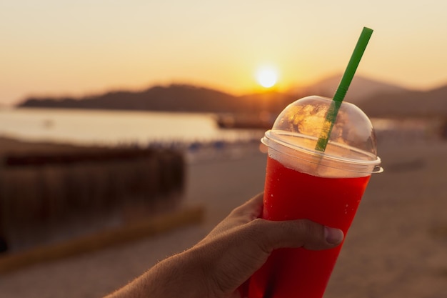 Detail einer nicht erkennbaren man39s-Hand, die bei Sonnenuntergang am Strand ein Glas matschiges Getränk im Vordergrund hält