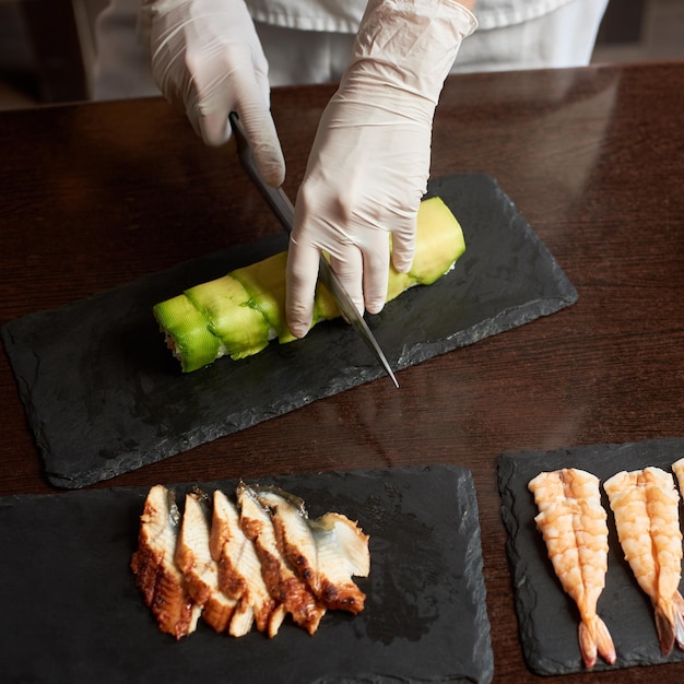 Detailansicht des Prozesses der Zubereitung von rollenden Sushi. Hände in Handschuhen Schneiderolle auf der schwarzen Steinplatte