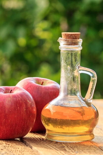 Detailansicht von Apfelessig in Glasflasche mit Kork und frischen roten Äpfeln auf alten Holzbrettern