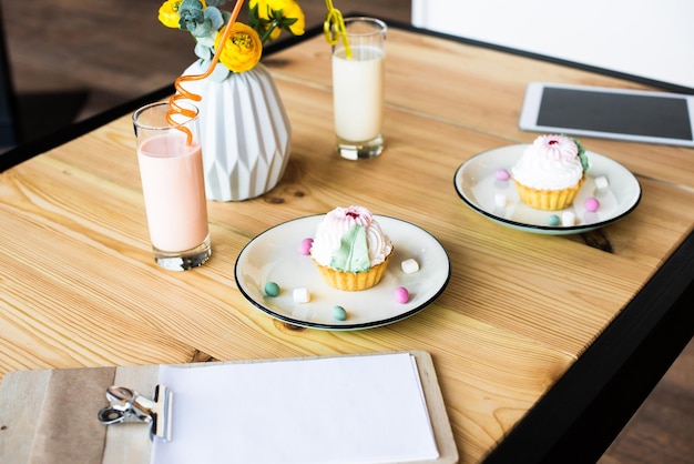 Detailansicht von leckeren Cupcakes aus der leeren Zwischenablage auf Tellern, Milchshakes und digitalem Tablet auf Holz