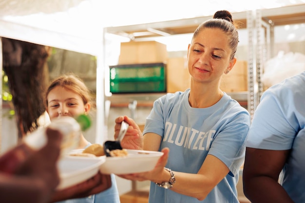 Detailliertes Bild, das eine kaukasische Wohltätigkeitsmitarbeiterin zeigt, die einer benachteiligten afroamerikanischen Person warme Mahlzeit serviert. Freiwillige Frau verteilt kostenloses Essen an hungrige und bedürftige Obdachlose.