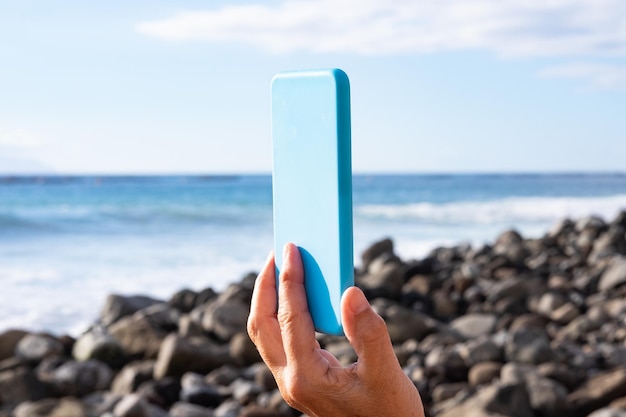 Detalhe da mão de uma mulher sênior na praia segurando um celular com capa azul clara Horizonte sobre o mar