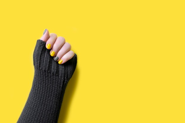 Detalhe da mão de uma mulher vestindo um suéter cinza com cores da moda iluminando as unhas de manicure amarela e cinza