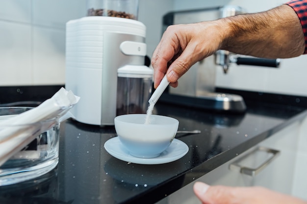 Detalhe das mãos de um homem despejando açúcar em uma xícara com café expresso