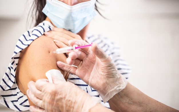 Detalhe de um médico aplicando a vacina Covid-19 por meio de injeção no braço de uma mulher usando máscara facial.