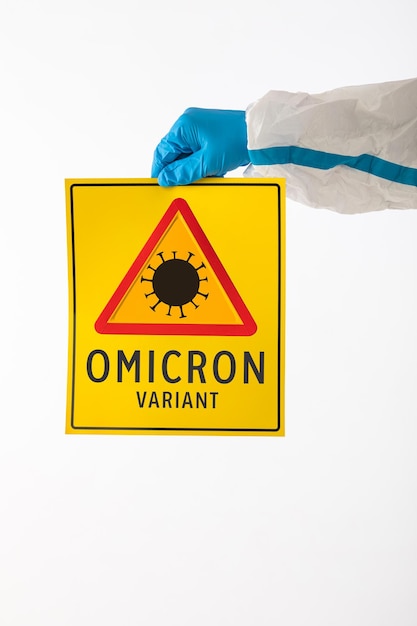 Detalhe do braço de uma enfermeira médica usando um EPI e luvas de látex com uma placa amarela com um símbolo de perigo COVID-19 que diz: 'Variante Omicron'. Coronavírus, pandemia e conceito de saúde.