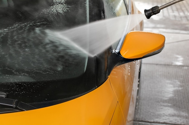 Detalhe na janela dianteira do carro amarelo escuro e no espelho traseiro sendo lavados com jato de água no lava-jato.