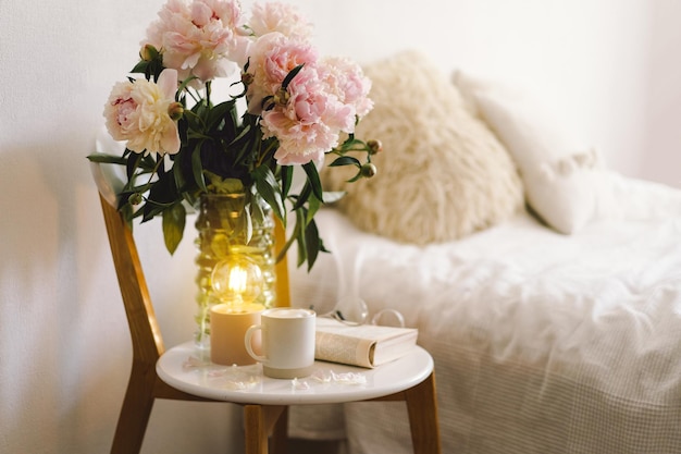 Detalhes da vida ainda no interior da sala de estar Livro aberto com copos xícara café e buquê flores peônias rosa brancas Leia e descanse Casa aconchegante