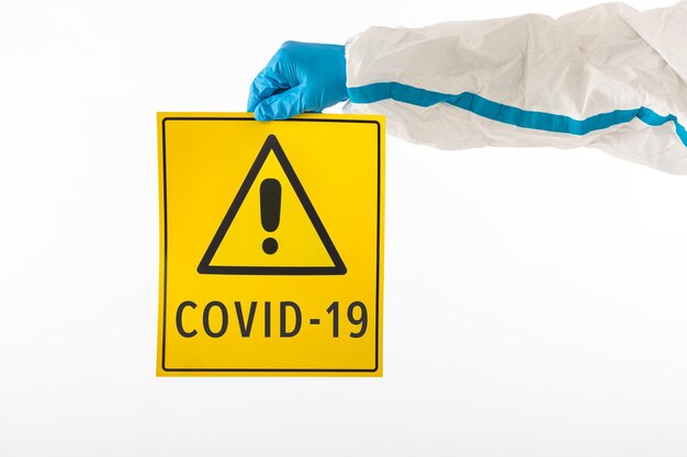 Detalle de un brazo de una enfermera médica con un EPP y guantes de látex con un cartel amarillo con un símbolo de peligro que dice: 'COVID-19'. Coronavirus, pandemia y concepto de salud.