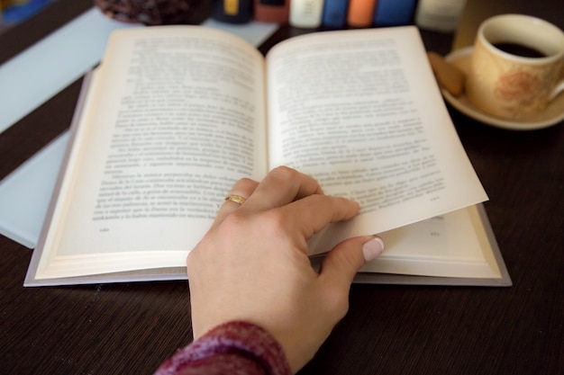 Detalle de la mano femenina que da vuelta a una página del libro en la mesa de madera oscura con más libros y una taza de café