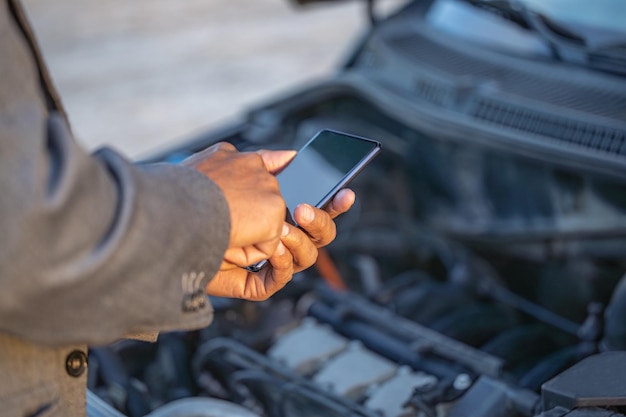 Detalle de las manos de un hombre latino con su teléfono móvil con su auto averiado al fondo