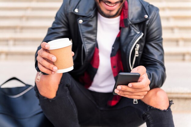 Detalle de las manos de un joven feliz irreconocible que consulta el teléfono con un café en la mano, concepto de tecnología y estilo de vida urbano