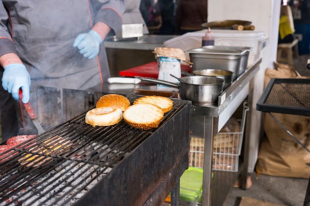 Detalle de una persona irreconocible cocinando hamburguesas en una parrilla de carbón caliente con especial atención a los panecillos tostados en la rejilla superior de la cocina del restaurante