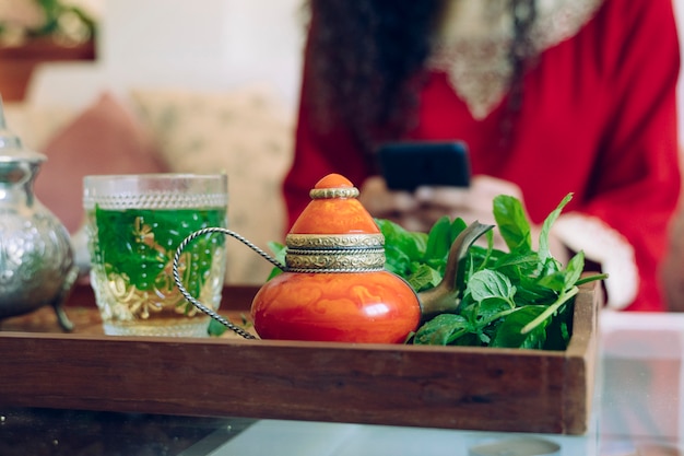 Detalle de té verde árabe en una bandeja y hermosa mujer joven con teléfono inteligente