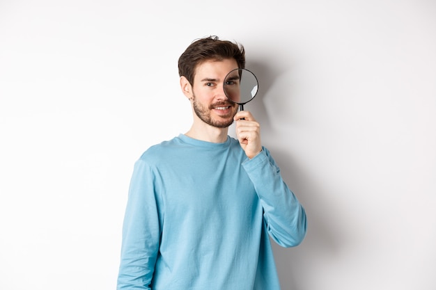 Foto detective joven sonriente mirando a través de la lupa, buscando oferta promocional, de pie sobre fondo blanco.