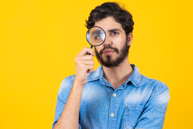 Detective masculino joven sosteniendo una lupa con el ojo derecho mientras está de pie frente a la cámara de forma aislada