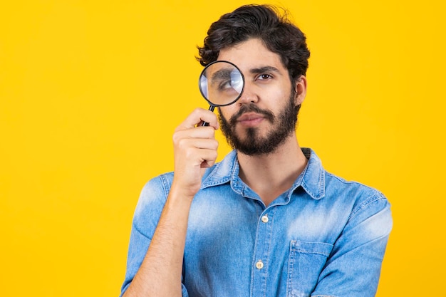 Detective masculino joven sosteniendo una lupa con el ojo derecho mientras está de pie frente a la cámara de forma aislada