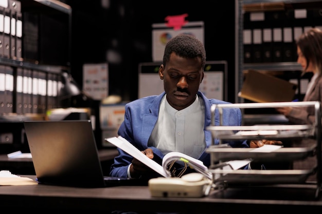 Detective privado leyendo archivos de criminología, buscando nuevas pistas para atrapar a un misterioso sospechoso. Oficial de policía afroamericano analizando pruebas delictivas trabajando horas extras en la sala de archivos