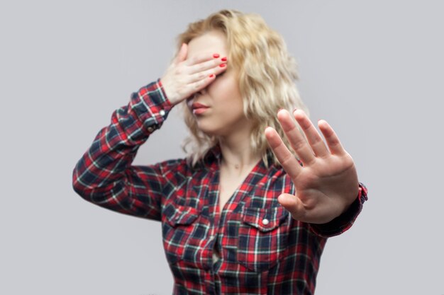 Detente, no quiero ver esto. Retrato de mujer joven rubia hermosa en camisa a cuadros roja casual de pie con gesto de parada y tapándose los ojos. tiro del estudio, aislado en fondo gris.