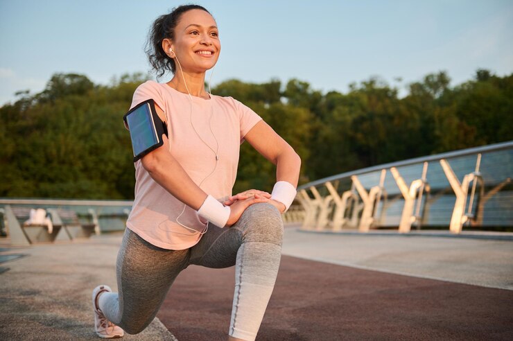Foto determinada desportista de meia idade com fones de ouvido e suporte para smartphone aproveitando o treino matinal ao nascer do sol, esticando as pernas, fazendo lunges, agachando antes de correr. atleta feminina malhando ao ar livre