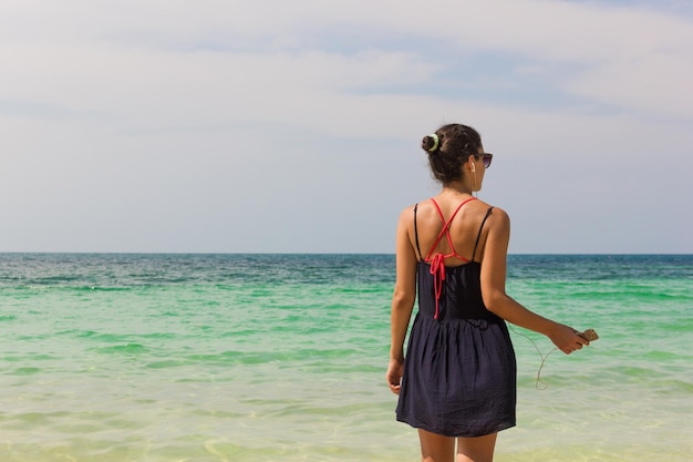 Detrás de una chica escuchando música frente al mar mientras sostiene el celular en un día soleado en la playa